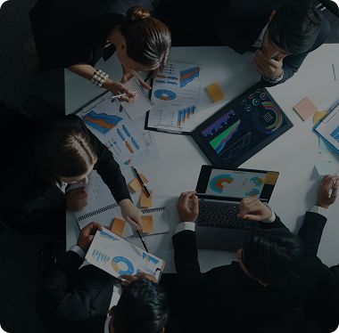 Overhead view of a team of financial advisors reviewing investment charts, graphs, and reports on paper and laptop screens around a table, representing PRG MFT's mission